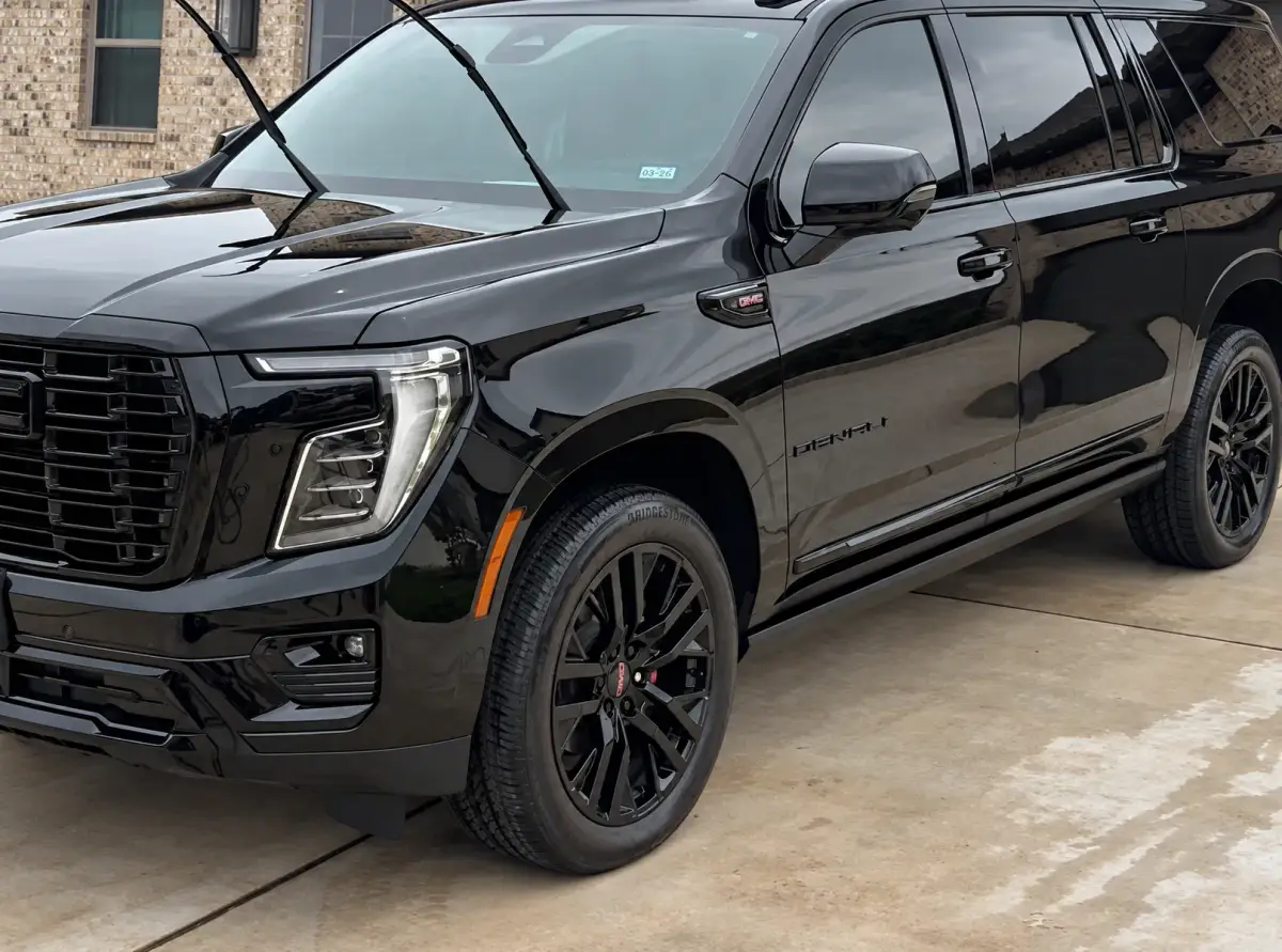 A glossy black SUV sits on a concrete driveway beside a brick house, with its windshield wipers raised and wet patches on the pavement.
